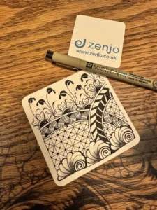 A completed black and white Zentangle tile resting on a wooden table next to a Sakura Pigma Micron pen and a Zenjo Tangle Club coaster.