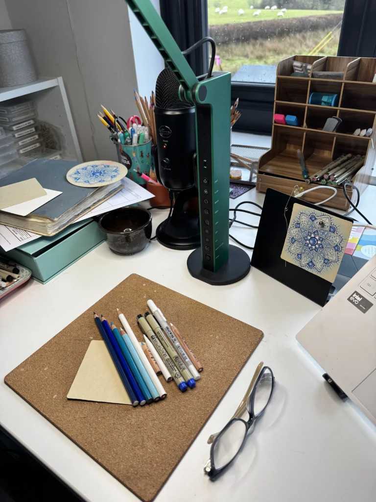 A calm workspace setup for Zentangle drawing, featuring a cork pad, Micron pens, blending pencils, a microphone, and a finished blue mandala tile.
